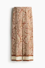 Pantalón de corte holgado en tejido dobby color marrón con estampado de hojas en color crema. Modelo de tiro alto con elástico en la parte posterior de la cintura y cierre oculto con corchete en un lado. Bolsillos laterales discretos y piernas amplias con pliegues adelante y atrás.