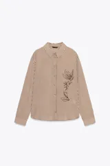Camisa a rayas verticales marrones y blancas, de corte recto, con cuello solapa y mangas largas con puños abotonados. Presenta bordado floral con abalorios en el frente.