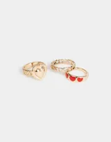Set de tres anillos dorados de metal. Uno con tres corazones rojos esmaltados, otro con un corazón liso y otro con textura.