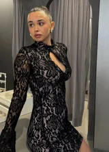 Vestido corto de encaje negro con forro color piel, cuello alto con abertura en el pecho y mangas largas acampanadas.