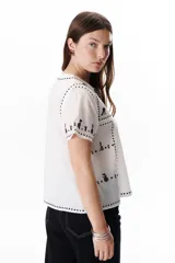 Blusa negra con bordados blancos de animales y motivos geométricos. Tiene cuello redondo con lazo y mangas cortas.