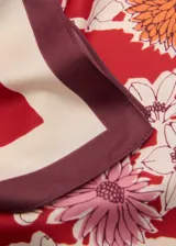 Pañuelo cuadrado con estampado de flores en tonos naranja, rosa y blanco sobre fondo rojo. Tiene un borde liso en color crema y un borde exterior en color borgoña.