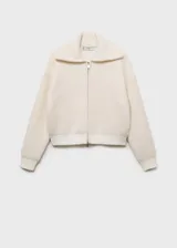 Abrigo corto color crudo de tejido bouclé con mezcla de lana. Tiene cuello de solapa ancha, cierre delantero con cremallera, manga larga con puños elásticos y terminaciones de canalé en contraste. Diseño oversize.