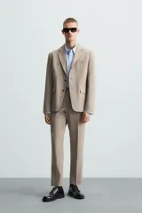 Pantalón de vestir para hombre, color beige claro con textura de tejido de lana. Presenta un corte regular fit, con pliegues marcados en el frente, bolsillos laterales y bolsillos traseros de doble vivo con botón.