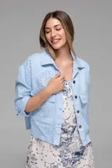 Campera corta color coral con cuello camisero, cierre frontal con botones, bolsillos de parche en el pecho y mangas largas con puños abotonados. Presenta calados tipo broderie en hombros, mangas y espalda.