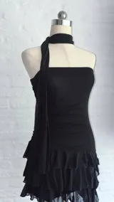 Vestido negro, sin mangas, con escote asimétrico y detalle de lazo o corbatín largo que cae desde el hombro. El cuerpo es ajustado y fruncido, y la falda corta tiene volados superpuestos.