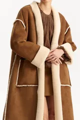 Abrigo largo de diseño envolvente, exterior de cuero gamuzado color marrón y forrería interior de corderito color beige. Presenta solapas anchas de corderito y puños también de corderito.