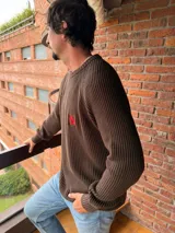 Sweater de tejido de punto color beige, con cuello redondo y manga raglán. Presenta un pequeño bordado en hilo rojo en el pecho.