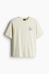 Remera color crema de corte holgado, con cuello redondo y estampado pequeño de Snoopy y la frase "Peanuts" en el pecho.