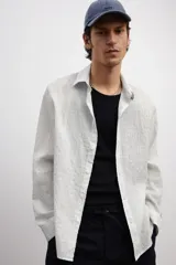 Camisa blanca de popelina de algodón texturizado, con cuello inglés, tapeta lisa, canesú con pliegue de caja en la espalda y mangas largas con abertura y cierre ajustable de botón en los puños. Corte holgado y bajo levemente redondeado.