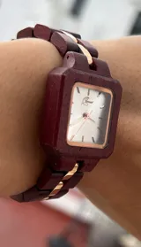 Reloj de pulsera con caja cuadrada y correa de eslabones de madera color borgoña. El dial es plateado con agujas y detalles dorados.