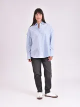 Camisa celeste de corte amplio, con cuello clásico, cierre frontal con botones, bolsillo en el pecho y mangas largas con detalle de botones en los puños.