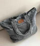 Bolso grande tipo totebag de jean azul, con asas de mano y asa larga para colgar. Se cierra mediante 1 botón de metal a presión y contiene 3 bolsillos exteriores.