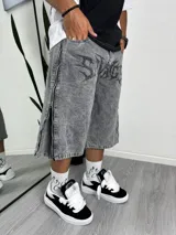 Bermuda de jean gris lavado, corte ancho (baggy), con estampado frontal en negro con tipografía tribal de la palabra "Savage". Presenta cierres decorativos en los laterales de las piernas.