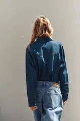 Camisa de denim azul con cuello solapa, manga larga, dos bolsillos delanteros con solapa y cierre frontal con botones a presión.