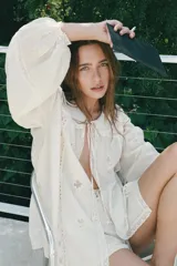 Camisa blanca de algodón con cuello bobo, mangas largas abullonadas con puños elásticos y cierre frontal con lazo. Presenta bordados florales y detalles de encaje.
