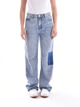 Jean de mezclilla azul claro con efecto acid wash, corte recto y tiro alto. Presenta un parche de mezclilla azul oscuro en el muslo derecho y un bolsillo utilitario en el muslo izquierdo. El ruedo está deshilachado.