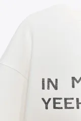 Sudadera corta color crudo de algodón, con cuello redondo, mangas largas y texto estampado en el frente que dice "IN MY YEEHAW ERA".