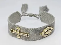 Pulsera tejida color gris con cierre ajustable y dijes dorados: una cruz con strass y un dije ovalado con la imagen de la Virgen María rodeada de strass.