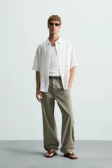 Camisa de hombre de manga corta, cuello solapa y corte relaxed fit. Tejido blanco con textura sutilmente arrugada o plisada. Cierre frontal de botonadura y bajo con aberturas laterales.