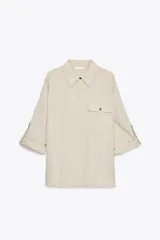 Camisa color beige de cuello solapa y manga larga con trabilla y botón para remangar. Tiene un bolsillo de plastrón en el delantero, bajo asimétrico y cierre frontal con botones ocultos por una solapa.