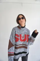 Sweater tejido de algodón con cuello alto, mangas largas y diseño con rayas horizontales en tonos marrón y negro. Estampa frontal con la palabra "SUR" en letras rojas.
