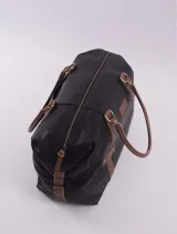 Bolso de viaje negro con detalles en cuero marrón, doble asa de mano y cierre superior con cremallera.