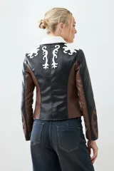 Chaqueta corta de cuero sintético color negro, marrón y blanco, con cuello de piel sintética blanca, cierre frontal con cremallera y bordados en las mangas.