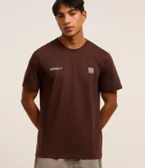 Remera marrón de algodón, corte clásico, con cuello redondo y manga corta. Presenta un pequeño estampado con lettering y un logo en el pecho.