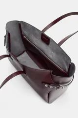 Bolso tipo shopper grande de cuero sintético color borgoña (vino tinto), con doble asa de mano y detalles de ojales metálicos plateados en los laterales. Incluye asa extraíble para llevar al hombro o cruzado.