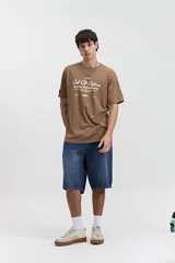 Remera marrón de manga corta con cuello redondo y estampado frontal con la frase "Out Of Office BUT STILL CHASING DREAM".