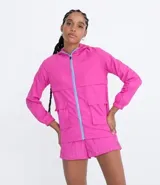 Campera rompeviento alargada color fucsia, con capucha, cierre frontal con cremallera color lila, bolsillos con solapa y puños elásticos.