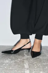 Zapato destalonado de piel negro, con punta fina y tacón kitten bajo y curvo. Posee una tira trasera con pieza elástica para ajuste en el talón.