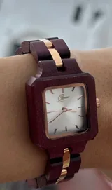 Reloj de pulsera con caja cuadrada y correa de eslabones de madera color borgoña. El dial es plateado con agujas y detalles dorados.
