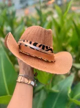 Sombrero de ala ancha tejido en hilo de coco y sintético, color marrón claro. La copa está cubierta con encaje naranja quemado con patrón floral. Tiene una cinta de cuero marrón oscuro y un aplique decorativo de hoja de gamuza con incrustaciones de pedrería.