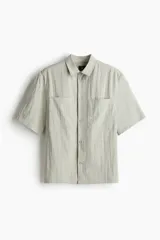 Camisa color verde claro con cuello inglés, tapeta lisa y canesú en la espalda. Modelo de manga corta con dos bolsillos superiores abiertos y bajo recto con aberturas laterales.
