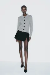 Chaqueta de punto gris con cuello solapa y manga larga. Tiene bolsillos delanteros de plastrón, rib en los acabados y cierre frontal con botones a presión en forma de lazo.