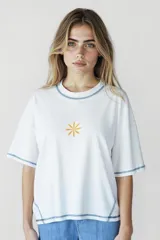 Remera blanca de corte holgado con cuello redondo y mangas cortas. Presenta un pequeño estampado de una flor amarilla en el centro del pecho y detalles de costura en azul en cuello, mangas y ruedo.