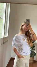 Remera blanca de algodón con cuello tipo bote y detalle de elástico en los hombros, creando un efecto de caída asimétrica.