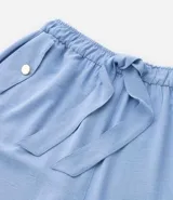 Pantalón de crepé celeste, modelo zanahoria, con elástico y lazo en la cintura. Presenta bolsillos traseros simples y bolsillos delanteros con solapas y botones metalizados.