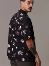 Camisa de manga corta con estampado de flores y hojas en blanco y gris sobre fondo negro. Tiene cuello clásico, cierre con botones y bolsillo de parche en el pecho.