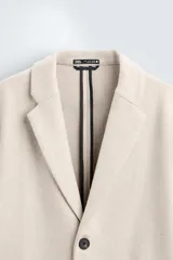 Abrigo straight fit color beige confeccionado en tejido con lana. Cuello con solapas de muesca, manga larga acabada en puño con botones, bolsillos de vivo en cadera, detalle de bolsillo interior y bajo con abertura en centro espalda. Cierre frontal de botonadura.