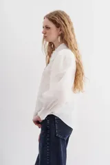 Camisa blanca de algodón con corte recto, cuello clásico y cierre frontal con botones. Presenta detalles de puntilla en cuello y cartera, mangas largas abullonadas con puños elastizados.