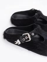 Zueco negro de pelo vacuno, estilo cerrado con abertura en forma de T en el empeine, asegurado con una tira de cuero y hebilla metálica. Incluye un dije metálico con forma de llama en la tira.