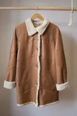 Abrigo gamulán marrón con cuello, solapa, puños y borde frontal de piel sintética blanca. Tiene cierre con botones y bolsillos laterales.