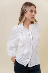 Camisa blanca de tela liviana con detalles de encaje y terminaciones caladas. Presenta cuello con leve volado, botonera frontal y mangas largas abullonadas con puños ajustados.