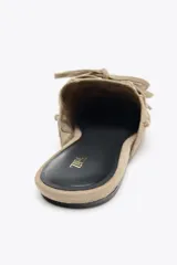 Zapato plano destalonado tipo mule, color beige, confeccionado en serraje. Presenta antifaz y lazo con borlas en la parte delantera.