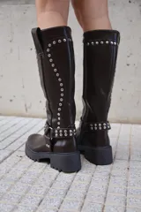 Bota alta de caña ancha color negro, estilo biker o motera, con suela track gruesa y taco bajo. Presenta apliques de tachas plateadas a lo largo de la caña y una correa con hebilla y argolla decorativa alrededor del tobillo.