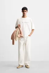 Camiseta blanca de corte holgado, cuello redondo y manga corta. Presenta un pequeño estampado de una estrella azul y un texto en negro en el frente.