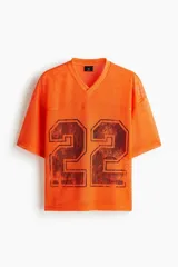 Remera de malla color naranja vibrante, estilo jersey de fútbol americano, con cuello en V y corte oversized. Presenta un estampado grande y desgastado del número "22" en color negro o marrón oscuro en el frente.